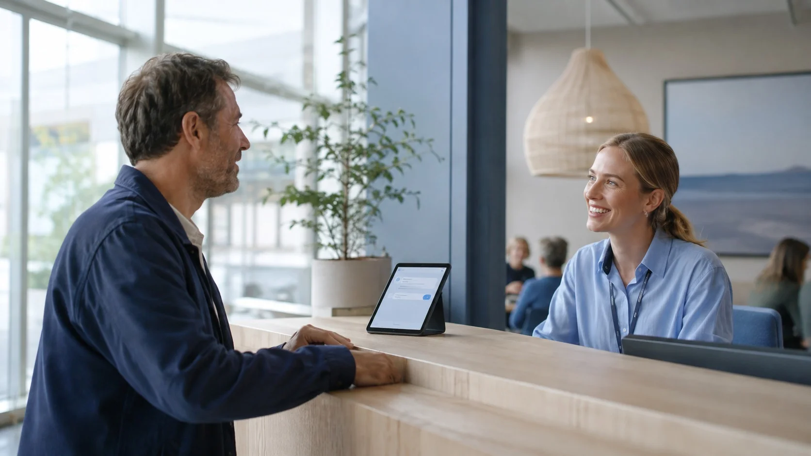 Kunde ved en skranke taler med en smilende medarbejder ved siden af en tablet